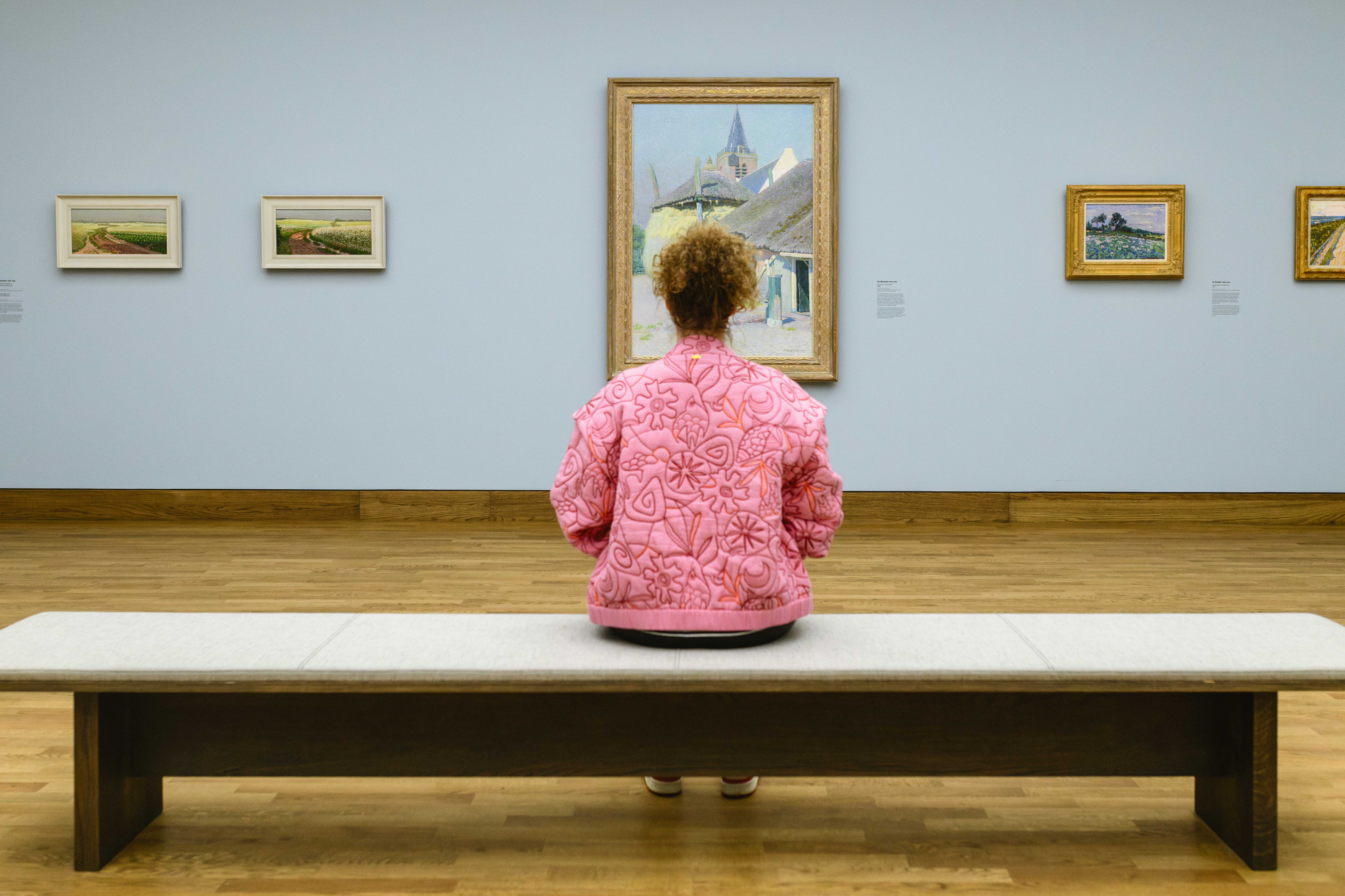 foto waarbij we tegen de rug van Margot Ros aankijken, zet zit op een lange bank in een museumzaal