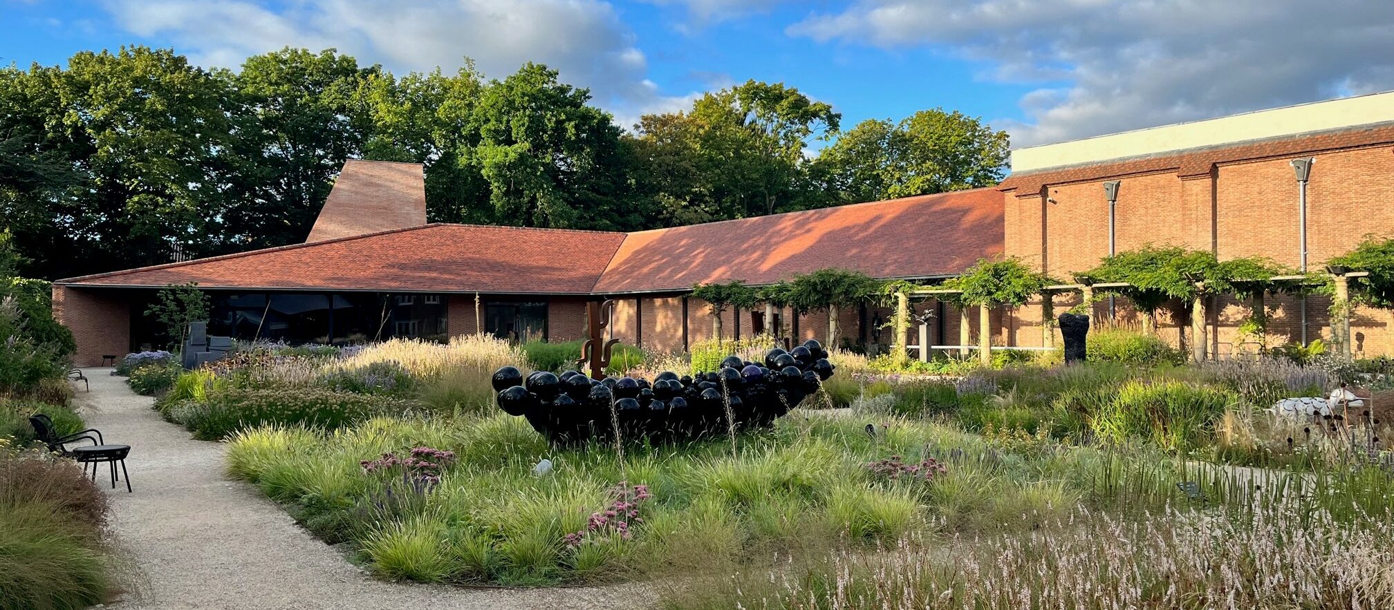 foto waarbij we tegen de rug van Margot Ros aankijken, zet zit op een lange bank in een museumzaal