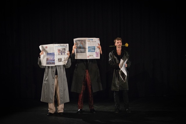 Drie mannen met lange regenjassen op een rij. Twee mannen hebben een krant voor hun gezicht. De derde heeft een bloem in zijn mond. Foto door Suzie Hagens