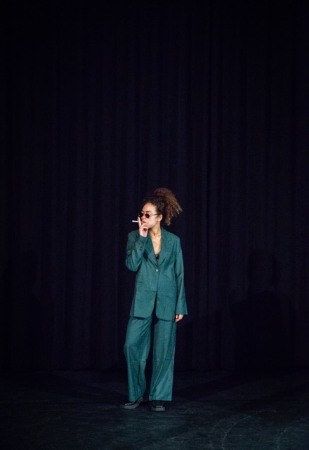 Vrouw in een groen pak met zonnebril op staat een sigaret te roken. De achtergrond is een zwart doek.
